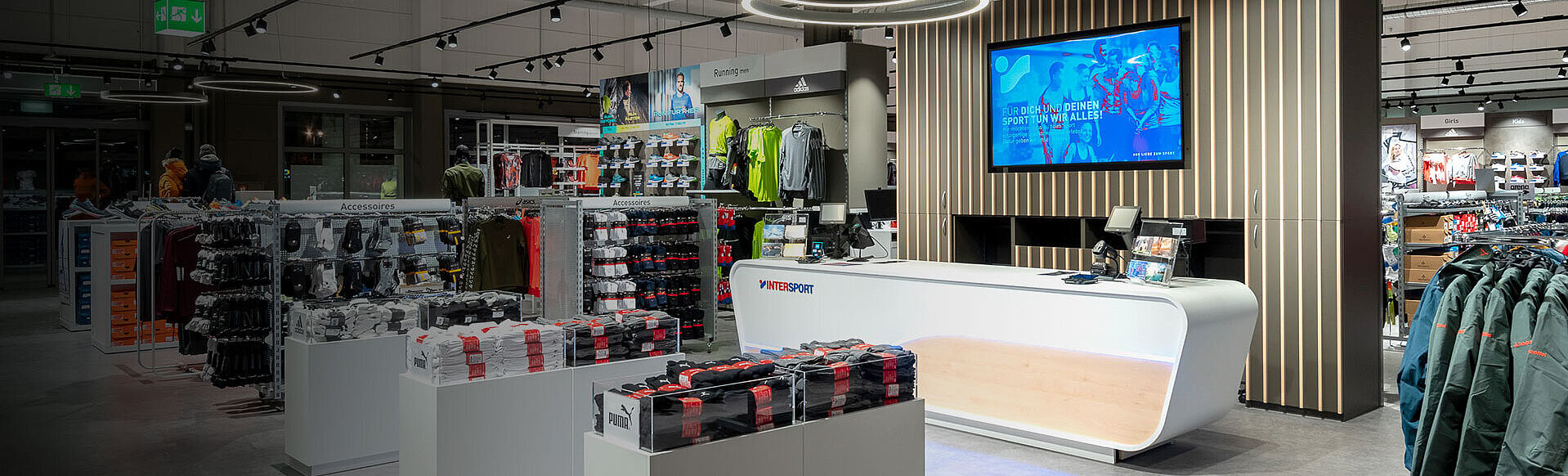 Verkaufsraum mit Regalen und Ständer für Sportartikel. Im Zentrum breite Kassentheke, dahinter großer Bildschirm für Werbung an der Wand.